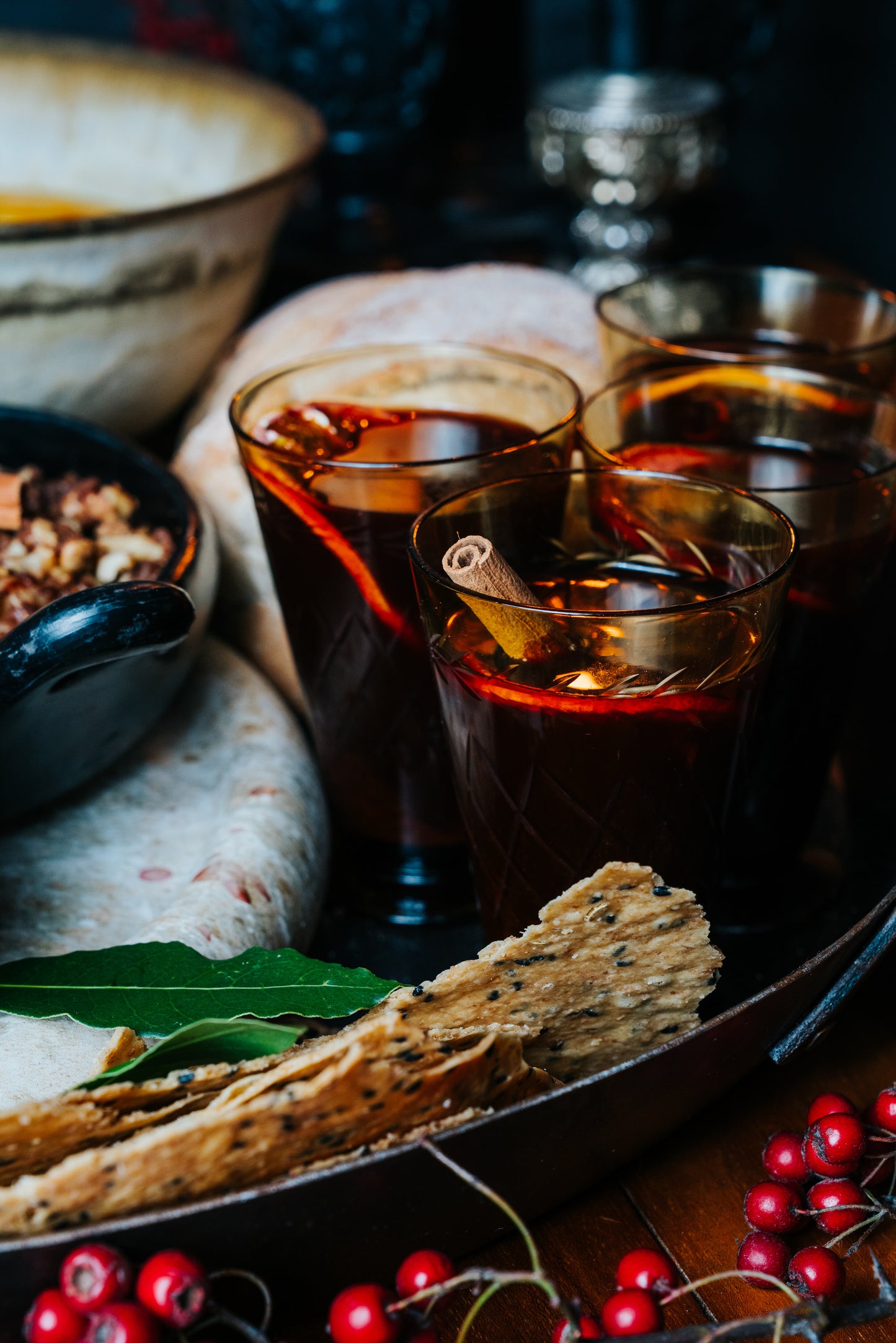 Three glasses of mulled wine with garnishes on a wooden tray with food items.